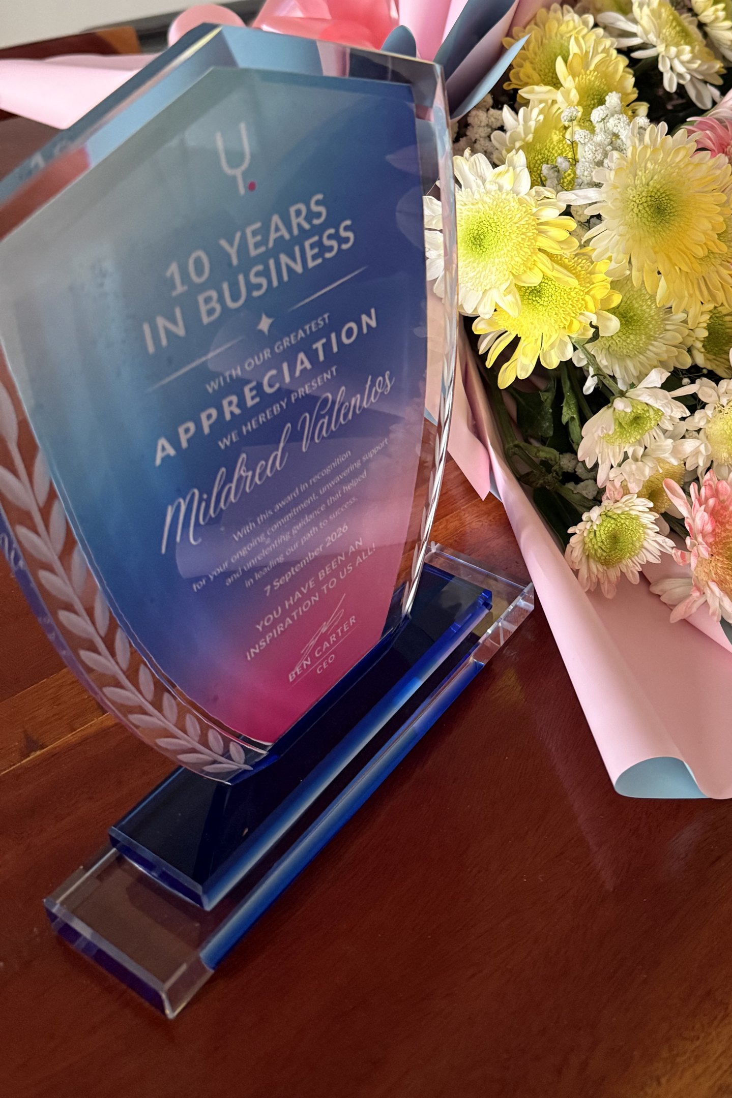 A glass award reading 10 years in business, with our greatest appreciation, Mildred Valentos, beside a bouquet of flowers